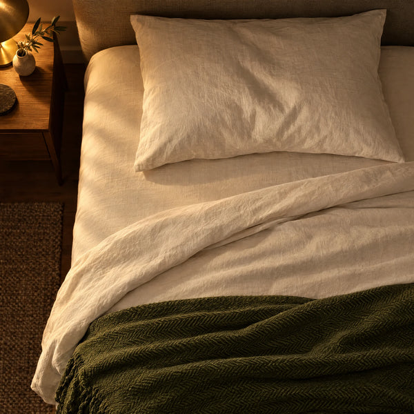 Cama tendida con sábanas de lino claro y una manta verde olivo, de noche